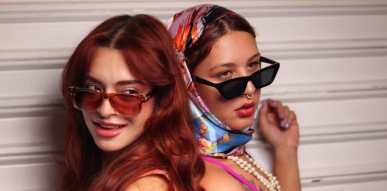 Two stylish young women, wearing fashionable sunglasses, posing in front of a textured background.