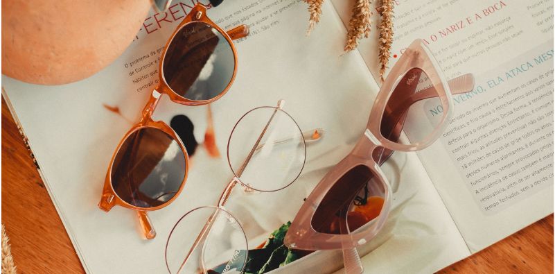 A flat-lay of orange club master style sunglasses, round eyewear and pink exaggerated cat-eye sunglasses on a magazine
